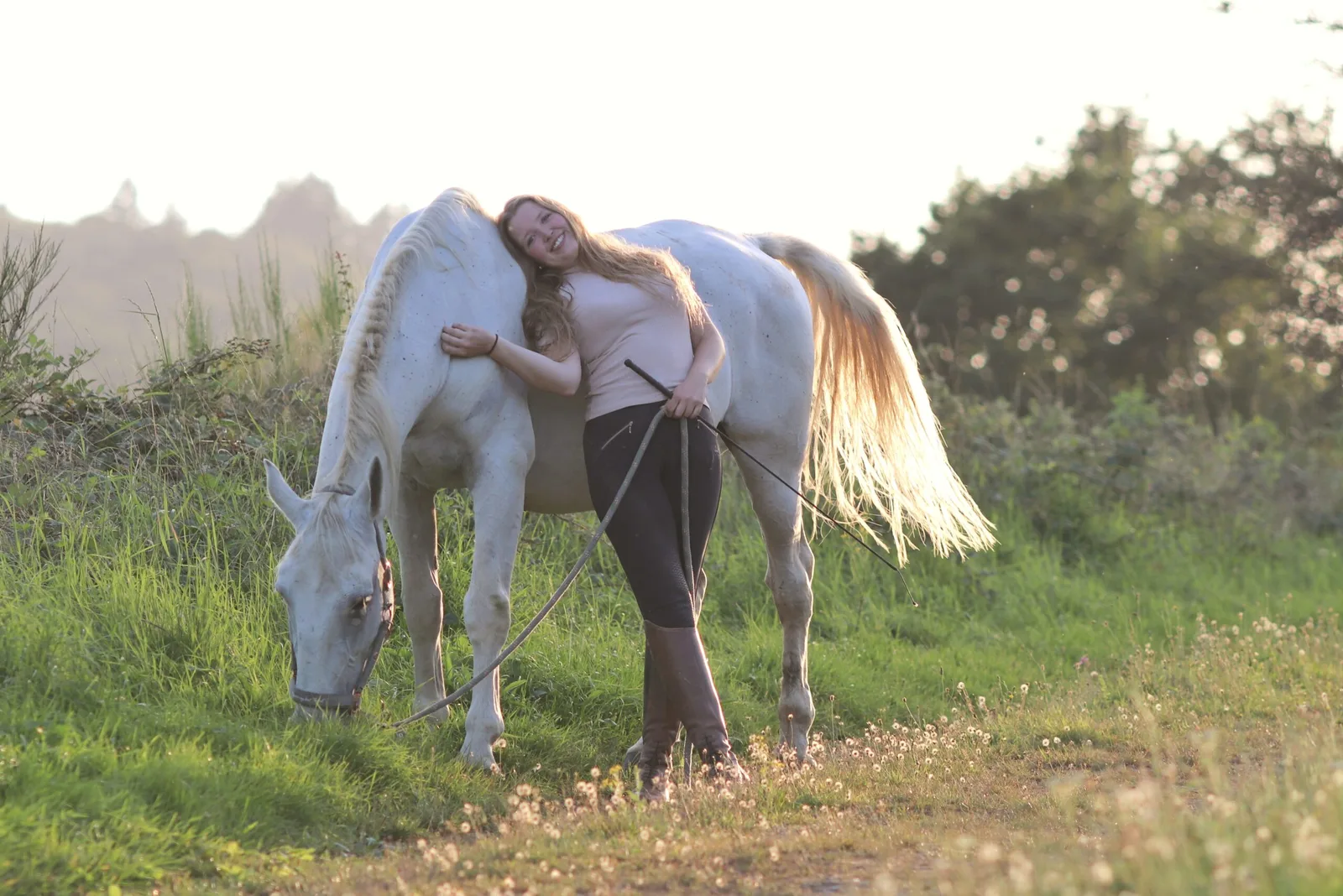 Frau mit weißem Pferd auf grüner Wiese im Gegenlicht, natürliche Pferdebeziehung, sanfte Stimmung
