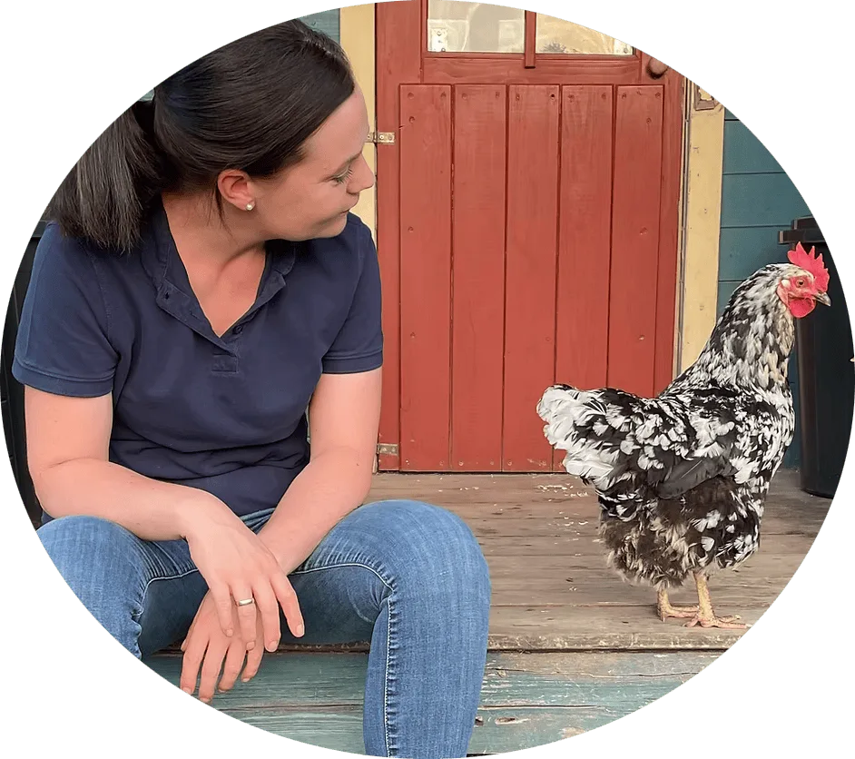 Frau mit dunklem Pferdeschwanz in dunkelblauem Polohemd sitzt vor roter Scheunentür neben Huhn