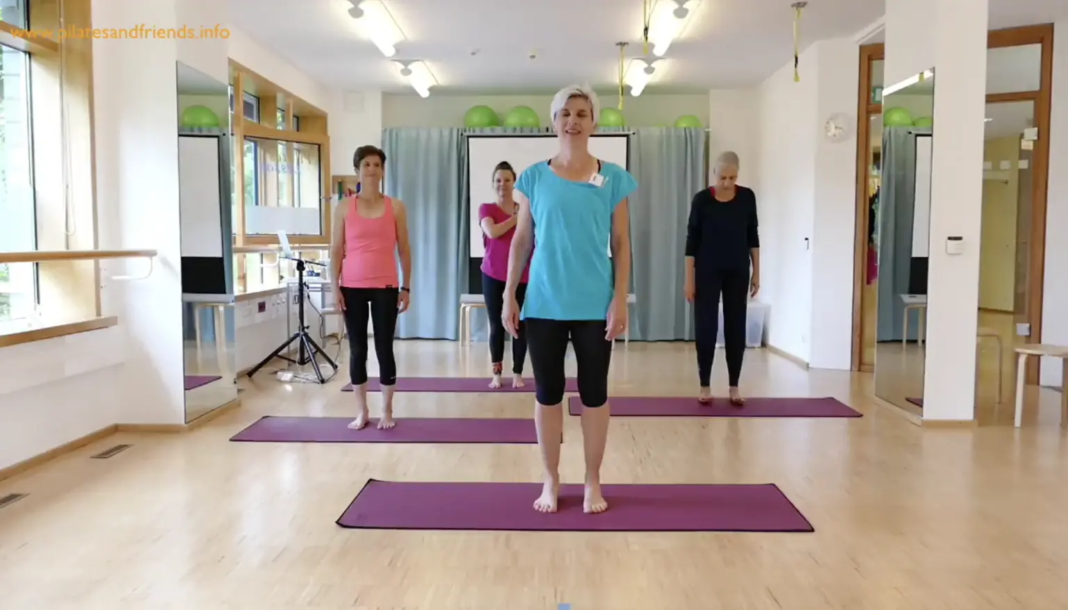 Pilates-Kursraum mit Instruktorin in blauem Shirt und mehreren Teilnehmern in verschiedenen Farben