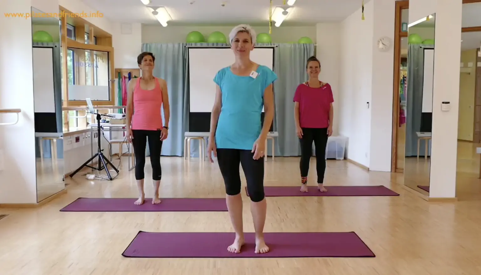 Fitnesstrainer in blauem Shirt führt Pilates-Kurs in Trainingsstudio vor, drei Teilnehmer auf violetten Matten
