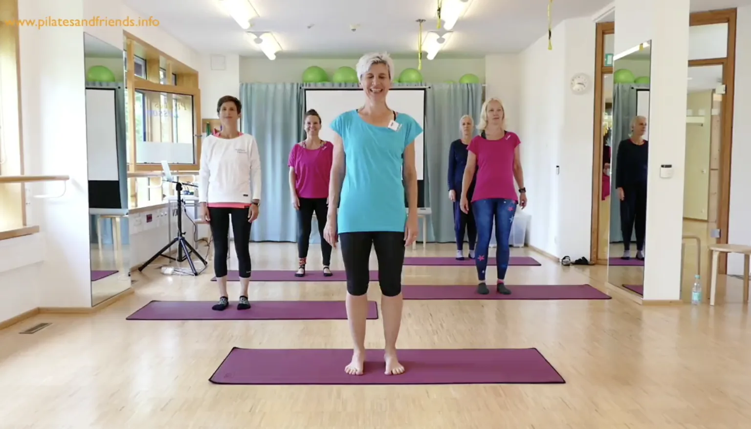 Pilates-Kursraum mit Instruktorin in blauem Shirt und mehreren Teilnehmern auf lila Matten