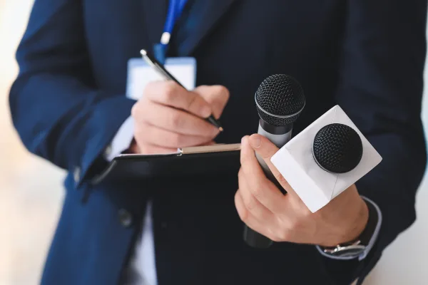 Journalist in dunklem Anzug hält zwei Mikrofone und Notizbuch für Interview