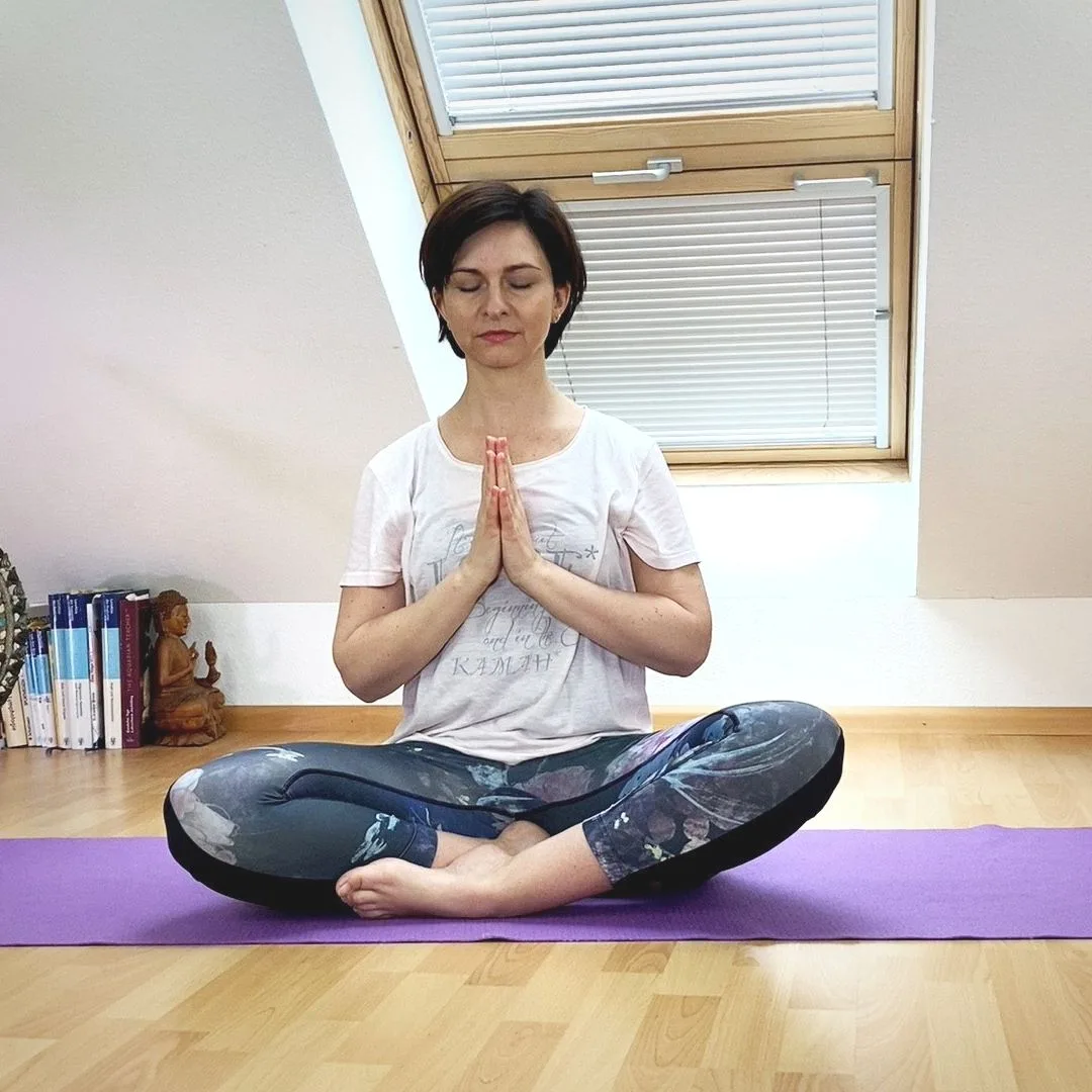 Frau in Meditationshaltung auf lila Yogamatte auf Holzboden, Gebetshaltung, helles Studio mit Fenster, Bücher und Deko sichtb