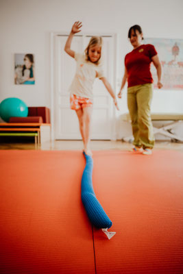 Kind auf rotem Trampolin balanciert mit erhobenen Armen auf blauer Balance-Rolle