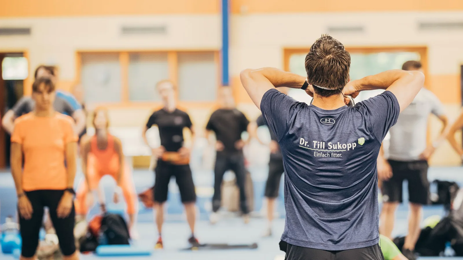Zurück zur Kraft – Videokurs für Beweglichkeit von Till Sukopp 14 Fitness-Trainer Till Sukopp unterrichtet eine Gruppe von Teilnehmern in einer Sporthalle mit orangefarbenen Elementen