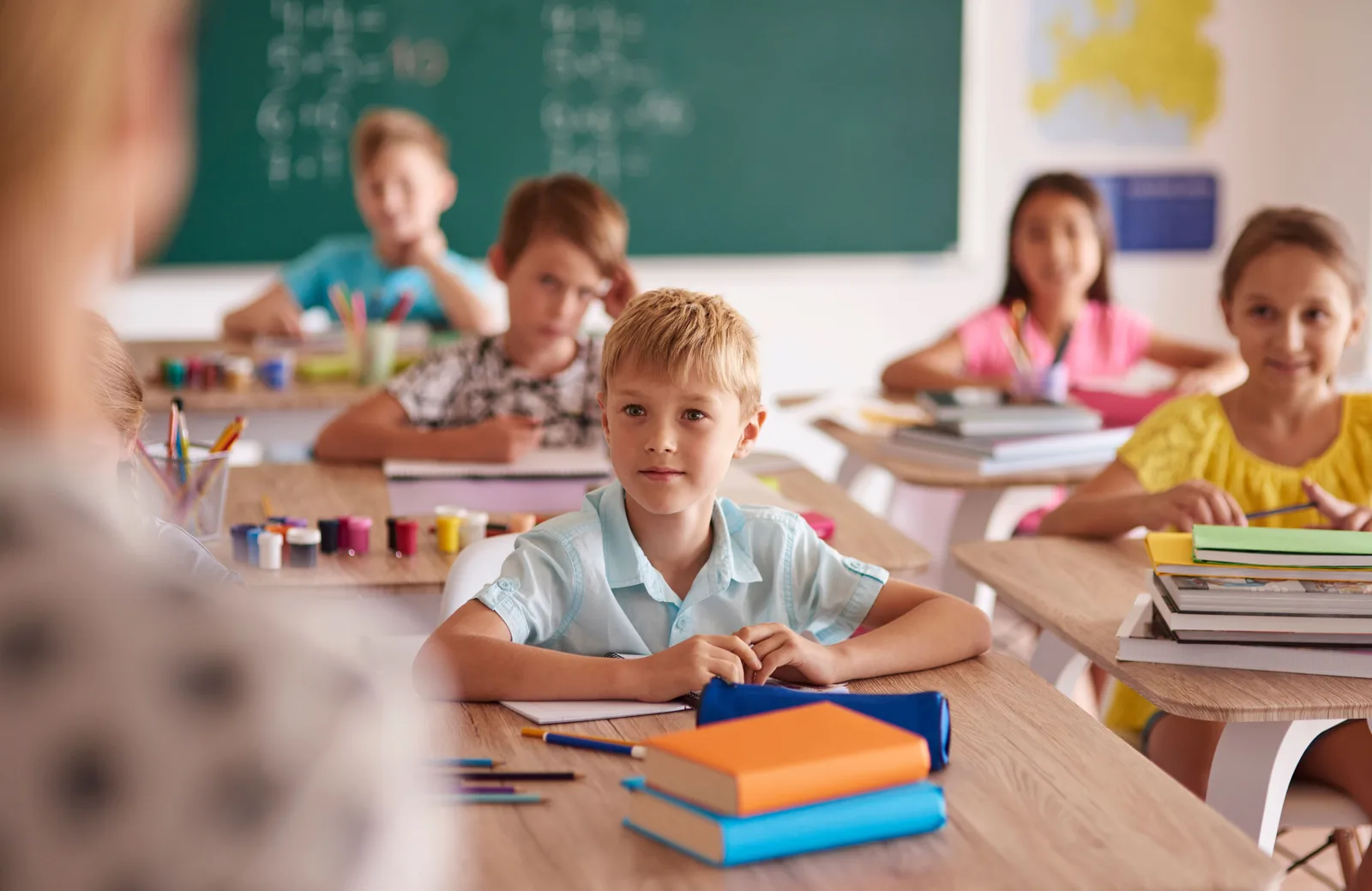 Klassenzimmer mit Schülern an Schultischen, Junge in hellblauem Hemd im Fokus, Tafel im Hintergrund