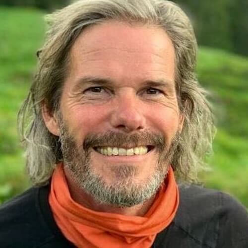 Porträt von Christian Meier mit langem grauem Haar, Bart und orangem Schal vor grünem Hintergrund