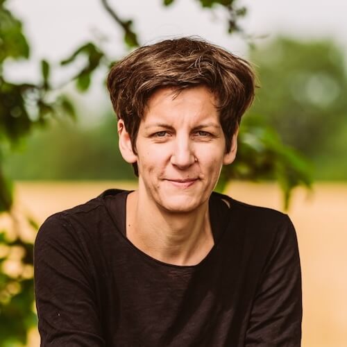 Porträt von Anne Kriesel mit kurzen braunen Haaren, schwarzem Shirt vor unscharfem grünem Hintergrund